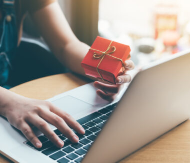 Woman's hand entering information into a laptop while holding a red gift box in her hand.